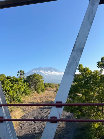 The image captures a scenic view of Mount Agung in Kubu, Indonesia, from what appears to be a bridge or elevated walkway. In the foreground, two red metal railings are visible, attached to white structural supports of the bridge. Beyond the railings, a dirt path with scattered rocks runs through dry grass and low vegetation. A dense line of green trees borders the path on the left, with a solitary palm tree standing tall amidst them. Further in the distance, the majestic Mount Agung is prominently featured, its peak partially shrouded by white clouds. The sky is a clear, vibrant blue, suggesting a bright, sunny day. The overall impression is one of natural beauty and tranquility.
