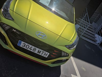 The image shows the front of a lime green Hyundai i10 N Line parked in a lot with a white line marking. In the background, a concrete staircase with a metal railing leads upwards. The license plate on the car reads "3048 MTH". The scene appears to be located in the city of Collado Villalba, Spain, possibly near an apartment building or commercial area. There are no people visible in the image. The lighting suggests it is daytime, with bright sunlight casting shadows.