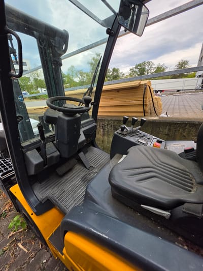 This image shows the interior of a yellow and black forklift parked outdoors in Zeppelinheim, Germany. The forklift is facing away from the camera. The steering wheel, control levers, and seat are visible. The environment appears to be an industrial or construction site, with stacks of lumber and a truck in the background. The sky is overcast.