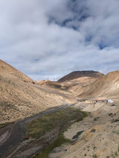The image depicts a wide, arid valley with a winding river flowing through it. The terrain is characterized by barren, sandy hills and slopes, with sparse patches of green vegetation along the riverbanks. In the mid-ground, a small cluster of buildings, appearing to be houses or shelters, is visible on a hillside. The sky is partly cloudy with a mix of white clouds and patches of blue. The location is identified as Pang, India, a high-altitude settlement. A small red object, possibly a tent or a vehicle, is situated near the river on the left side of the frame. The overall scene conveys a sense of remote, rugged landscape.