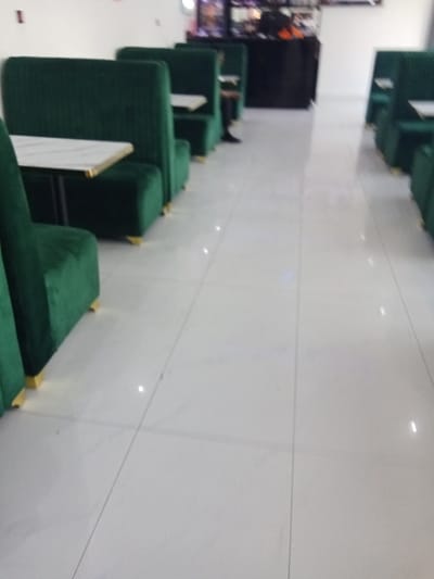 The image shows an empty restaurant with a row of green booths on the left side of the frame and a similar row of booths on the right side of the frame. All the booths are empty. The floor is white tile. There is a black counter in the background with a person sitting on a stool at the counter. There is a table with a white surface and metal legs in the foreground. The image is likely taken at a restaurant or cafe in Ujevwu, Nigeria. There are no other people present in the image.