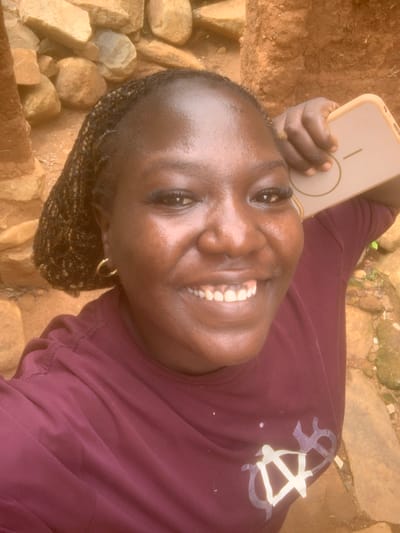 A close-up selfie shows a Black woman with braided hair and gold hoop earrings smiling while holding a light-colored phone to her head. She is wearing a dark maroon t-shirt with a white graphic design on the left side of her chest. The background consists of rocks and dirt, suggesting an outdoor setting, possibly in Zhwarki Mile 10, Nigeria. The lighting is bright, highlighting the sheen on her skin.