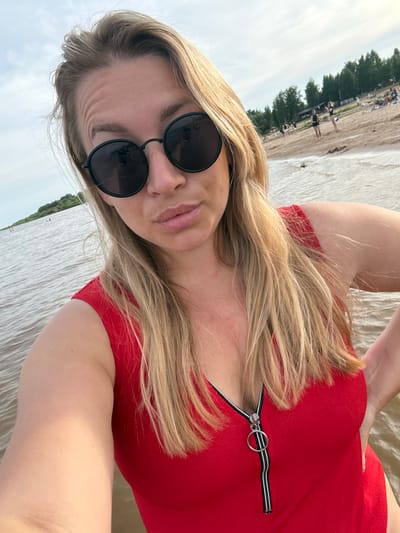 This is a selfie of a young woman at Ondvor, Russia. She has blonde hair and is wearing sunglasses and a red top with a zipper. She is on a beach with trees in the background. The weather is cloudy.
