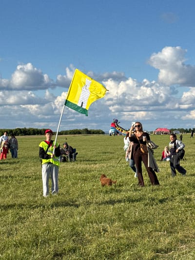 In Bolgar, Russia, on a grassy field under a partly cloudy sky, a young person wearing a high-visibility vest and a red cap stands holding a yellow flag with a drawing of a white castle on it. To their right, a woman in dark clothing takes a photo with her phone, holding a cup in her other hand. Behind her, other people are visible, including a woman with a backpack and another woman sitting on the ground. A small, brown dog is in the foreground, and a colorful inflatable structure is visible in the background. The overall scene suggests an outdoor event or festival.