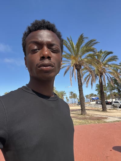 A young Black man is taking a selfie outdoors on a sunny day. He is looking towards the camera with his eyes closed and a slight smile on his lips. He is wearing a black t-shirt and a silver chain necklace. The background shows palm trees, a clear blue sky, and a red brick walkway. There is also a white vehicle and a blue sign with some text visible in the distance. The overall mood is relaxed and happy, suggesting a casual outing on a warm day. The scene appears to be in a coastal area, possibly a promenade or park, given the presence of palm trees and the brickwork. The time of day is likely late morning or early afternoon, as indicated by the bright sunlight and clear sky.