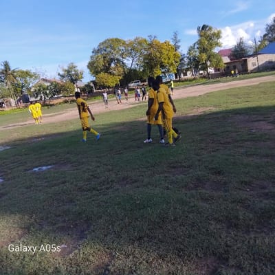 The image depicts a group of young men playing soccer on a grassy field in Nungwi, Tanzania, located at coordinates -5.726 latitude and 39.301 longitude. The men are wearing yellow and black soccer uniforms. Several of them are standing around a goalpost in the middle of the field. There are several trees and a few houses in the background. The sky is blue with a few white clouds. A soccer ball is visible in the bottom left of the image. The image was taken with a Samsung Galaxy A05s.