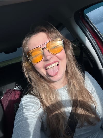 A young woman with long, light brown hair is inside a car, wearing sunglasses and sticking out her tongue. She is wearing a gray long-sleeved shirt and a seatbelt. The car window has water droplets on it, suggesting it may have been raining. The image provides no information about the location in Verkhnie Iziverki, Russia.