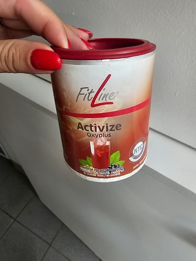 This media file is a close-up, angled shot of a person's hand holding a cylindrical container of "FitLine Activize Oxyplus." The container is predominantly white with red and orange branding. The text "FitLine" is prominently displayed in a stylized red font, followed by "Activize Oxyplus." There is also a circular seal with "NTC" and "Nutrient Transport Concept" visible. The label also indicates it's a "Natural Health Product / Produit de santé naturel" and "in powder form / en poudre." The background is blurred, showing a light-colored wall and a tiled surface, suggesting an indoor setting. The person's fingernails are painted a bright red. The lighting appears to be neutral, indicating it could be daytime indoors or under artificial light. The overall impression is that of a product being presented or held.