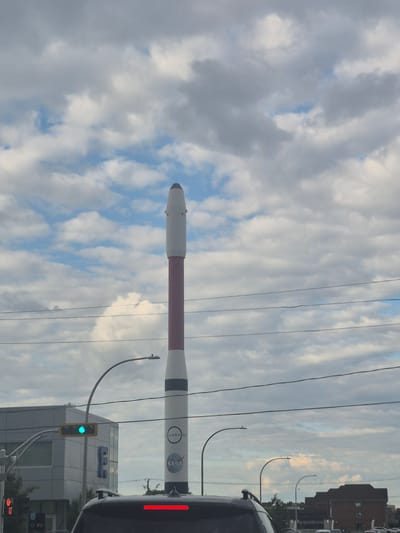 The image shows a view of a tall, rocket-shaped sculpture with "NASA" and "COSMOS" written on it, situated in Laval, Canada. The sculpture is white with a red band in the middle and has a black tip. It is positioned in front of a cloudy sky. To the left of the sculpture, a building with a light gray facade and blue lettering is visible. A traffic light with a green signal is hanging from an arched pole. Power lines are strung across the scene. In the foreground, the top of a black vehicle with a red reflection on its roof is visible, suggesting the photo was taken from inside another vehicle. The overall environment appears to be urban, with a focus on the unique sculpture.