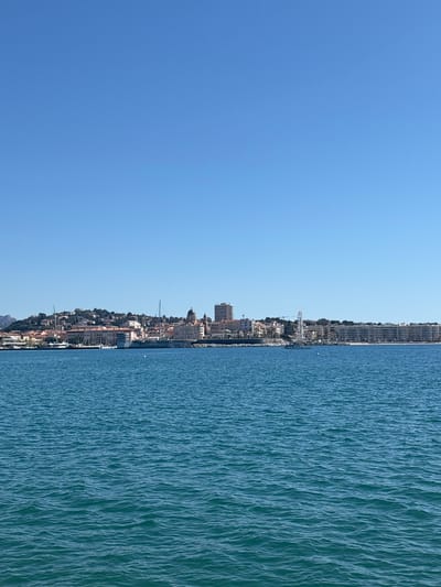 The image shows a view of Fréjus, France, from the sea. The right half of the image is the Mediterranean sea, with small wave patterns. The left half shows the city of Fréjus, a coastline crowded with buildings, including some high-rise apartments. A large Ferris wheel is visible. The image is oriented vertically, with the sky on the left, the city in the middle, and the sea on the right. The weather appears to be sunny.