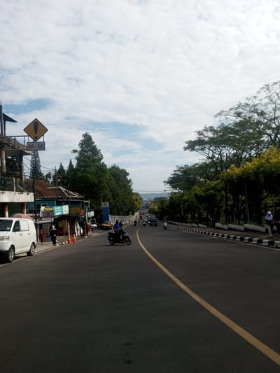 The image depicts a street scene in what appears to be a suburban or semi-rural area. The foreground shows a wide asphalt road with a yellow center line. A motorcycle is traveling on the road, and other vehicles are further down the road in the distance. On the left side of the road, there are shops and a white van parked. People are walking along the sidewalk in front of the shops. A road sign with an exclamation point and writing in a foreign language is visible. On the right side of the road, there is a line of trees and a black and white striped curb. The sky is partly cloudy. Based on the architecture, road markings, and signage, it's unlikely the location is Zeyathiri District, Myanmar. The image features elements more common in Southeast Asian countries like Indonesia or Malaysia.