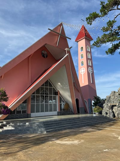 The image shows a pink church with a tall, slender tower in Đa Thọ, Vietnam. The church features a distinctive triangular roof with a white trim and a large cross at its peak. The tower, also pink, has a red roof and is adorned with arched windows and a prominent cross symbol. String lights are draped across the scene, suggesting a festive or decorative element. In the foreground, there are wide stone steps leading up to the church entrance, which is comprised of large, metal-framed glass doors. To the right of the church, a rocky formation with carved figures and text is visible. The ground in front of the church is a paved courtyard with some scattered pink flower petals. The sky is a clear blue with some wispy white clouds. The overall scene conveys a bright and serene atmosphere.