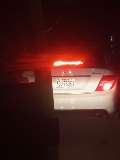 This media file captures the rear view of a white Mercedes-Benz sedan at night, likely in Dabore, Nigeria. The car's brake lights are illuminated, casting a strong red glow that dominates the scene and causes some lens flare. The license plate is clearly visible and reads "ABJ-768KJ", with "ABUJA" printed above it. The "4 MATIC" designation is also visible on the right side of the trunk. The setting appears to be on a road, possibly in traffic, given the illuminated brake lights. The overall mood is dark and somewhat obscured, with shadows and light from the car's taillights. The image quality is moderate, with some graininess due to the low light conditions. There is also a reflection of the car's interior on the rear window or possibly on the license plate. The context of Dabore, Nigeria, suggests this image was taken in that specific region.