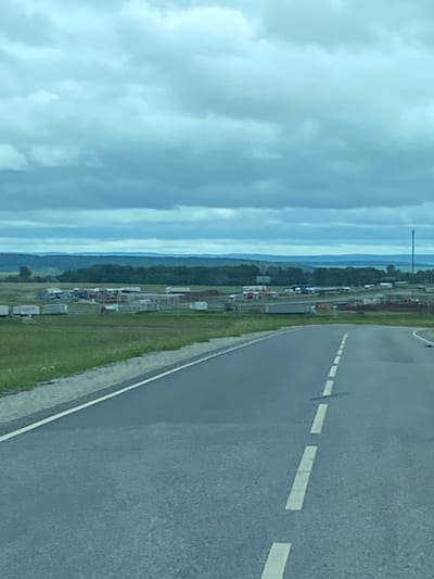 The image shows a rural landscape with a road in the foreground. The road has white dashed lines indicating lanes. Beyond the road, there is a grassy field dotted with various trailers, some appearing to be storage units, and what looks like a construction or industrial area with multiple structures and machinery. In the background, there is a line of trees and rolling hills under a cloudy sky. The scene is overcast and appears to be a day with low visibility due to the clouds. The location is identified as Kliuchiki, Russia. There are no people visible in the image.