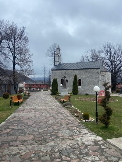 The image shows a stone path leading to a stone building in Kolašin, Montenegro. A statue stands in front of the building. There are bare trees on either side of the path, and benches are placed along it. The sky is overcast.