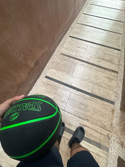 A high-angle, medium shot depicts a person holding a black and green Spalding basketball while standing on a textured stone walkway. The walkway features alternating light-colored rectangular stones and darker linear stone strips, bordered by small, loose gravel. A dark brown surface is visible to the left. The person is wearing dark-colored shorts and slides. The location cannot be determined as Mérida, Mexico without additional visual or contextual clues.