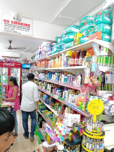 The image shows the interior of a store in Oxel, India. Aisles are lined with shelves stocked with a variety of products, including diapers, beverages (Coca-Cola), snacks, and personal care items. A man in a white shirt and jeans stands in one of the aisles. A woman in a pink floral dress is bending over. A "No Smoking" sign is visible on the wall. A display of Clipper lighters is present.