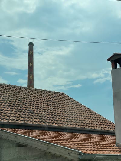 The image captures an outdoor daytime scene dominated by an industrial chimney and multiple rooftops against a cloudy sky. A tall, slender, reddish-brown brick chimney stands centrally, featuring vertically stacked Cyrillic letters painted in alternating yellow and red: 'К', 'Ъ', 'Р', 'Н', 'А', 'Р', 'Е', which spell "КЪРНАРЕ" (Karnare). This identifies the location as Karnare, Bulgaria. Below the chimney, several layers of terracotta-tiled rooftops with visible gutters are present, indicating residential or other buildings. On the far right, a partial view of a light grey, rectangular chimney or vent structure with a darker opening and a reddish cap is visible. A single dark utility wire extends horizontally across the upper portion of the image, positioned in front of the industrial chimney. The sky above is partly cloudy with patches of blue. No people or specific actions are depicted.