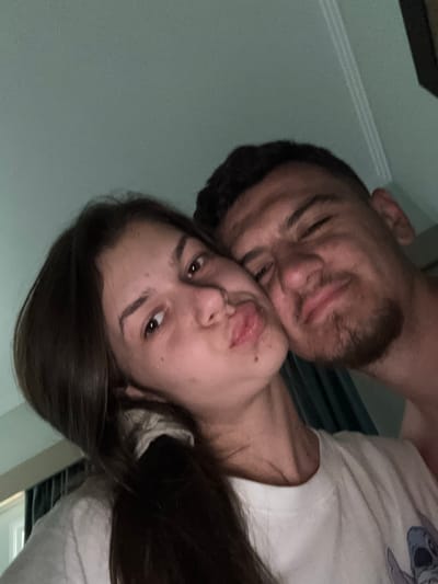 An intimate, close-up, and rotated selfie-style photograph captures two individuals, likely a couple, in a dimly lit indoor setting. The image primarily focuses on their faces, conveying a moment of affection and closeness.
On the left, a man with short dark hair, a beard, and a mustache is visible. His eyes appear closed or mostly closed, and his mouth is slightly open, suggesting a gentle nuzzle or kiss. His head is tilted back, partially obscured by the woman.
Resting against the man, on the right side of the frame, is the face of a woman with long, dark hair. Her dark eyes are open and looking slightly towards the camera, and her lips are slightly parted. She is wearing a light-colored t-shirt, upon which a blue and white cartoon character resembling Stitch from Disney's Lilo & Stitch is partially visible on her upper chest and shoulder area.
The overall mood is tender and affectionate. The background is largely dark and out of focus, indicating an indoor environment, possibly a bedroom or living space. Hints of a light-colored wall with horizontal molding lines are visible on the far left, and dark blinds or curtains can be vaguely discerned on the right. The low light conditions suggest the photograph was taken in the evening or at night. No specific textual elements are visible beyond the character on the shirt, and no direct cues to the Selçuk, Turkey location are present within the image.