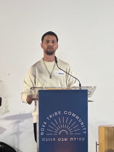 A male individual stands centered behind a clear acrylic and dark blue podium, positioned indoors against a light-colored wall in Kadima-Zoran, Israel. The man has short, dark hair and stubble, wearing a light cream-colored long-sleeved sweater, a silver-colored chain with a rectangular pendant, and a white name tag pinned to his right chest, displaying "Elkaha." His hands rest on the podium's clear surface. A black gooseneck microphone extends from the podium towards him. The dark blue front panel of the podium features white text: "NOVA TRIBE COMMUNITY" in an arc above a radiating sun graphic, and "קהילת שבט הנובה" below. The wall behind him shows faint dark discoloration on the lower left. A partial view of a golden object is visible to the right of the podium, and a dark object is on the floor to the left.