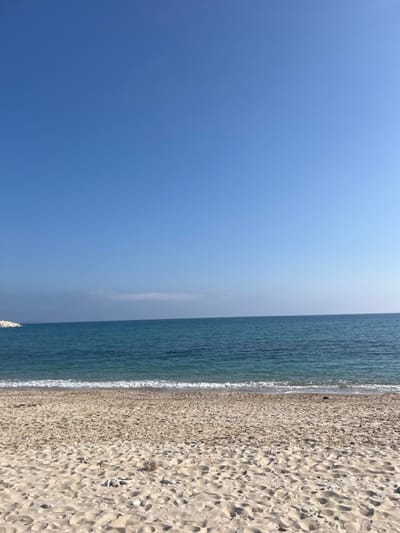 The image shows a wide, sandy beach in Limenaria, Greece, at coordinates 40.626, 24.571. The beach is empty, with no people visible. There are a few footprints in the sand, and a small rocky outcropping visible in the distance. The water is a clear blue and the sky is cloudless. There is a small wave crashing onto the shore in the foreground. The image is taken from a low angle, looking out towards the sea.