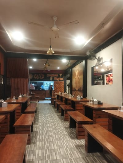 The image depicts the interior of a coffee shop and restaurant in Bandarbireuen, Indonesia. The scene is illuminated by overhead lights and features rows of wooden tables and benches, indicating a casual dining environment. A sign in the background reads "WD Coffee & Resto," confirming the establishment's identity. The restaurant appears to be empty or sparsely populated, with no prominent human activity captured. The decor includes wooden paneling on the walls and a large mural depicting an architectural landmark, possibly the Taj Mahal, adding an artistic touch. A ceiling fan is visible, suggesting a warm climate. The overall impression is that of a clean, functional, and inviting space designed for customers to enjoy food and beverages.