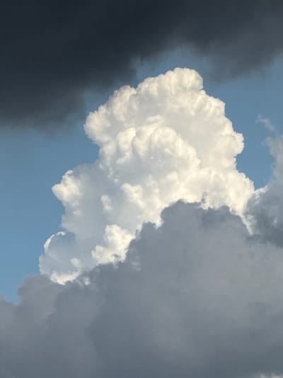 The image depicts a sky filled with dramatic clouds. A large, bright white cumulus cloud dominates the center, its voluminous form catching the sunlight and displaying intricate textures. To the left and upper portion of the frame, darker, gray storm clouds loom, creating a stark contrast with the bright cloud. The sky is a vibrant blue peeking through in some areas. The bottom portion of the image is filled with heavier, darker gray clouds, suggesting a storm might be approaching or passing. There are no people, structures, or discernible landmarks in this image, making its location in Đa Lộc, Vietnam, impossible to determine from the visual evidence alone. The scene is a natural phenomenon of cloud formations against a blue sky.