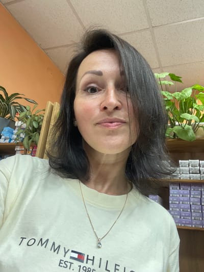 A woman with dark hair and fair skin is pictured from the chest up, looking directly at the camera. She is wearing a light yellow t-shirt with the Tommy Hilfiger logo and "EST. 1985" printed on it. A delicate gold necklace with a teardrop-shaped pendant is visible. Her hair is dark brown and falls just below her shoulders, with some strands framing her face. The background suggests an indoor setting, possibly a retail store, with shelves stocked with small boxes, likely hair dye or similar products, and some potted plants. The walls are painted a light orange color. The image does not provide information about the specific location within Kumyoni, Russia.