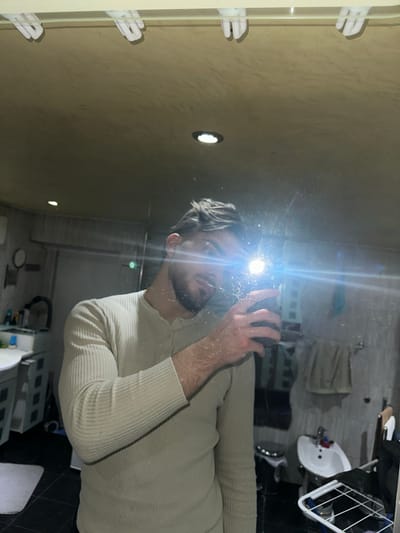 A mirrored selfie shot in what appears to be a bathroom. The reflection shows a man with short, styled hair and a beard, wearing a long-sleeved shirt. The background indicates a modern bathroom setting including a sink and cabinets. The image does not allow for any determination of the location in Rafz, Switzerland.