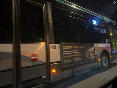 The image shows the side of a bus at night. The bus has the number "14" displayed near the top, along with the "ilede France" logo. An advertisement for "GODOT & FILS" is visible, stating that they are recognized experts with advisors in Paris and the Île-de-France region. The ad mentions "Or & Argent" and "Achat & Vente." A red circle with a horizontal line is near the bus door.