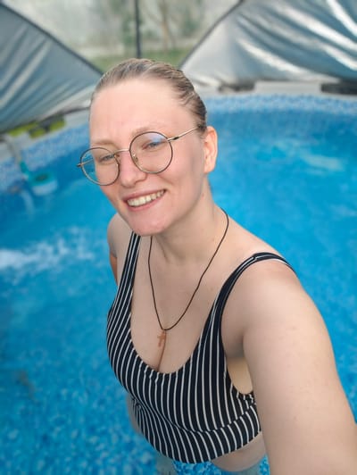 A young Caucasian woman with blonde hair, wearing round glasses and a black and white striped swimsuit, smiles at the camera while standing in a blue swimming pool. A cross necklace hangs around her neck. The pool is covered by a large, light-colored tarp, and the surrounding environment is blurred. The image was taken in Koledino, Russia.