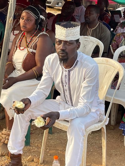 An outdoor scene captures a group of people gathered, likely for an event or social occasion, under what appears to be bright daylight. The ground is dry and earthy, typical of an outdoor setting.
In the foreground, a dark-skinned man is seated on a white plastic chair, looking directly towards the viewer. He wears a white, long-sleeved tunic with intricate dark embroidery around the neckline and chest, paired with white pants. A distinct white embroidered cap adorns his head. He has a dark beard and short dark hair. In both hands, he holds pieces of a light-colored, soft-looking food, possibly a cooked root vegetable like yam, suggesting he is eating or about to eat.
To the man's left (viewer's right, if the image were upright), a dark-skinned woman is also seated, her gaze directed downwards and slightly away from the camera. She is adorned in traditional attire, featuring a light-colored top with a brown, textured section across her chest, and multiple strands of vibrant bead necklaces in shades of red, green, white, and brown. Gold-colored bangles are visible on her left wrist.
Behind these two central figures, several other individuals are visible, mostly seated on similar white plastic chairs. One man directly behind the central man wears an olive green or khaki shirt and appears to be resting or looking downwards. Another person in the background wears a white t-shirt with partially visible text that seems to read "...AND YOU" or similar, though it's largely obscured.
A clear plastic water bottle with an orange cap is visible on the ground to the right side of the man, near his feet. The overall atmosphere appears casual and communal.