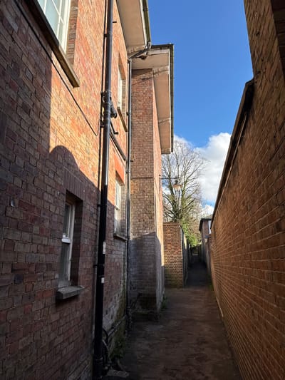 The image depicts a view looking upwards through a narrow alleyway in Dacorum, United Kingdom. The alley is enclosed by brick buildings on either side, with pipes running along the building facades. A patch of blue sky and clouds is visible above, along with a glimpse of a tree. The scene is captured with a wide-angle lens, resulting in a distorted perspective.