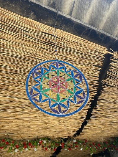 An overhead, close-up, low-angle shot captures a "Flower of Life" stained-glass suncatcher hanging from what appears to be a temporary thatched roof structure. The suncatcher is circular, framed by blue glass, and features a geometric pattern of petal-like shapes filled with rainbow-colored glass segments. It hangs by a thin string, from which a small, blue bead is also suspended. The thatched roof, made of tightly packed, light-brown reeds, forms the primary background. The reeds are oriented horizontally and are slightly irregular, suggesting a handmade or rustic construction. Above the reeds, a dark, rough surface is visible, possibly a roofing material or an overhang, with a grey, corrugated material above that. Along the bottom of the frame, a garland of artificial red and white flowers with green foliage is strung across the thatched structure, adding a touch of decoration. The scene is illuminated by natural light, casting subtle shadows and highlighting the colors of the suncatcher. The context of Olhão, Portugal, suggests this scene might be part of a traditional Sukkah decoration during the Jewish holiday of Sukkot, or potentially part of a local festival or market stall, given the common use of thatched roofs for temporary structures in the region.
