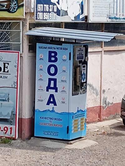 The image captures a water vending machine in Yukariyuz, Uzbekistan. The machine, colored blue and white, features the word "ВОДА" ("water" in Russian) prominently displayed, along with health-related icons and the slogan "КАЧЕСТВО ВОДЫ = КАЧЕСТВО ЖИЗНИ" ("water quality = quality of life"). It is situated next to a building with advertisements above and on its left side. The vending machine is protected by a corrugated metal roof. A car's headlight is partially visible on the right side of the image. The street in front of the machine is unpaved and dusty.