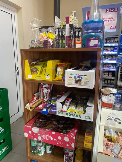 This image captures a section of an indoor retail setting, likely a convenience store or kiosk, located in Konstantinovo, Bulgaria. The scene is dominated by a three-tiered wooden display shelf unit in the foreground, packed with various consumer goods, primarily snacks, beverages, and confectionery items. No individuals are visible. On the top surface of the wooden shelf unit, from left to right, are: a clear cellophane-wrapped gift package tied with twine, containing small figurines; a dark purple patterned vase holding yellow flowers adorned with a pink ribbon; a stack of disposable paper coffee cups; several slim, black-packaged "ZERO" branded items in flavors like Eucalyptus and Mint; an orange Tic Tac box; a large clear plastic water pitcher with a blue lid, filled with water and topped with several inverted clear plastic disposable cups; a teal-colored plastic item with a sticker displaying "3.02лв / 5.92лв" (Bulgarian Leva, indicating a price); and a small, white decorative ornament resembling a feather or angel. Behind these items, a black vertical pipe is visible. The top shelf of the wooden unit contains two yellow cardboard display boxes on the left, each filled with individually wrapped "Меденка Желание" (Medenka Zhelanie - Honey Cake Desire) branded snacks featuring a cartoon donut. Visible prices are "0.50лв / 0.89лв". To the right, a white cardboard box labeled "Бишкотека синя слива" (Biskoteka Sini Sliva - Plum Biscuits), featuring a picture of plums, contains wrapped biscuits. A price of "3.00лв / 5.88лв" is partially visible. Other wrapped snacks are present behind these main displays. The middle shelf is filled with an assortment of individually wrapped chocolate bars and candies. Prominent brands include Toblerone (gold packaging), Kinder Bueno (red and white packaging), Oreo (blue packaging), and Milka (purple packaging). A white cardboard display box with green text "BOYANA СУХИ ПАСТИ" (BOYANA Sukhi Pasti - Dry Pastries) holds numerous individually wrapped pastries. The bottom shelf features a large red cardboard display box with white polka dots, branded "LUKAS," containing many individually wrapped items, likely chocolate bars or wafers. Beneath this box and to its right, several beverage bottles are visible, including a green cola bottle, two bottles of "ALOE" drink, a yellow bottle (possibly juice), and a dark-colored bottle. To the right of the "LUKAS" box, a stack of small yellow boxes labeled "MEGA CHIP" is present. Further to the right, a white cardboard box from "ЗАХАРНИ ЗАВОДИ" (Zaharni Zavodi - Sugar Factories)