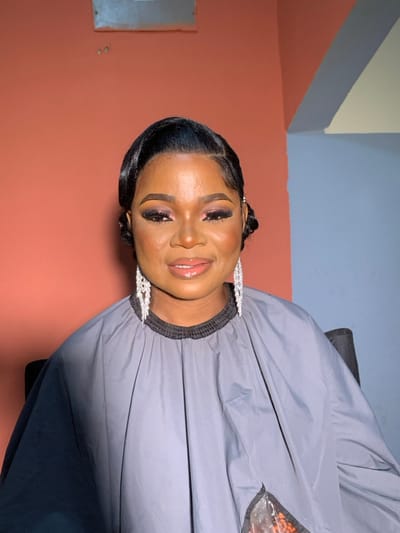 An adult female is centrally depicted in an indoor setting within the city of Garaku, Nigeria. She possesses dark skin, dark hair styled in sleek waves with a side bun detail, and expertly applied makeup including dark eyeshadow, eyeliner, and sculpted eyebrows, with glossy lips. She is wearing prominent, multi-tiered silver-toned chandelier earrings. Her torso is covered by a grey smock with a black elasticated collar, partially obscuring a dark chair. A translucent bag containing small orange items rests on the lower right section of the smock. The background consists of a wall painted reddish-orange on the left and light blue on the right, separated by an interior corner. An indistinct rectangular object and two small dark marks are present on the upper left wall. The subject maintains a subtle smile and faces directly forward.