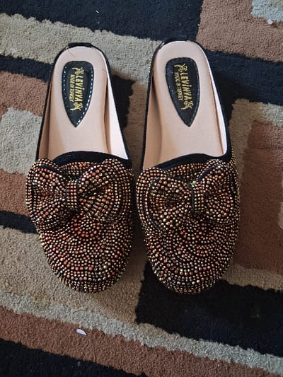 A pair of black, open-heeled women's slippers featuring densely applied rose gold-toned glitter studs on the vamp, forming a prominent bow-like structure, are positioned on a striped carpet. The slippers' insoles are light tan and display "LEVINNA MADE IN TURKEY" in black script. The carpet exhibits horizontal bands of black, light beige, and brown. A small, irregular white speck is visible on the light beige stripe of the carpet, towards the lower left. No individuals or actions are present. The scene is located in Gyadi Gyadi Arewa, Nigeria.