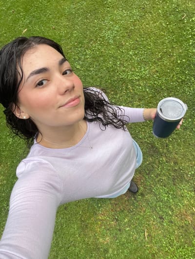 A high-angle photograph captures a young adult female standing outdoors on green grass. She possesses dark, wet, shoulder-length curly hair and a fair complexion. Her attire consists of a light purple long-sleeved top, light blue denim pants, and dark footwear. Accessories include a white pearl stud earring in her left ear and a thin silver chain necklace with a small, light-colored pendant. She holds a dark blue cylindrical thermal tumbler with a translucent lid in her right hand. Her gaze is directed towards the camera. The background consists entirely of green grass, appearing uniform and potentially damp. This scene is located in Huasca de Ocampo, Mexico.