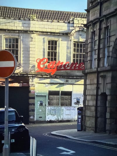The image depicts a building in Barnsley, United Kingdom, featuring a prominent red "Elite" sign. The building appears to be old, with multiple windows and a somewhat dilapidated facade. Next to it is an adjacent building that has been graffitied and appears dilapidated. On the right is a parking meter next to a road. A dark-colored car is parked on the side of the road. A red and white 'no entry' road sign is present in the foreground. The photograph is oriented at an unusual angle.