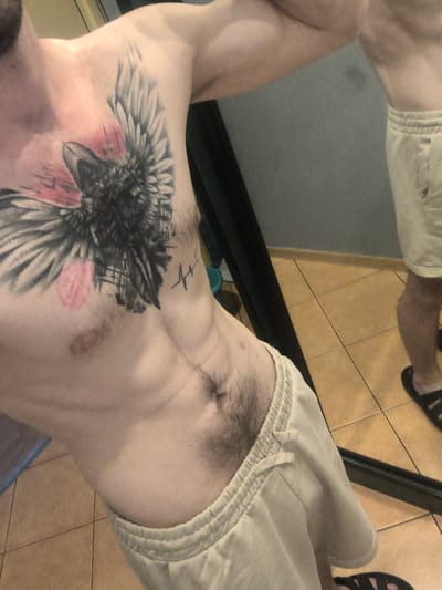 This is a medium-close-up, eye-level shot of a man's torso and upper thighs, with his right arm extended out of frame. The man has a tattoo of a raven with outstretched wings on his chest and collarbone, with pink and black coloring. He has a defined abdominal area with some body hair. He is wearing light-colored, drawstring shorts. The man is standing in what appears to be a bathroom or dressing area, as indicated by the tiled floor and a large mirror reflecting his lower body and legs. The man is barefoot. No specific location in Grójec, Poland can be identified from the image.