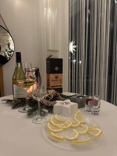A table is decorated with festive elements. There's a bottle of "Saint Martin" Chardonnay, two wine glasses, shot glasses, and a plate of sliced lemons. A candle holder in the shape of a star and a box with "Kazakstan" written on it add to the arrangement. Pine cones, ornaments, and greenery contribute to the ambiance. A string curtain is in the background, with a reflection of Christmas tree lights visible in a mirror.