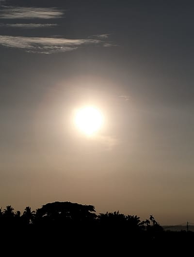 The image shows a sunset over Uleegajah, Indonesia. The sun is a bright, glowing orb in the center of the frame, casting a warm light across the sky. The sky is a mix of orange, yellow, and gray, with some wispy clouds visible. In the foreground, there is a silhouette of trees and palm trees, with a distant view of rolling hills and a telephone pole. The overall impression is one of peace and tranquility.