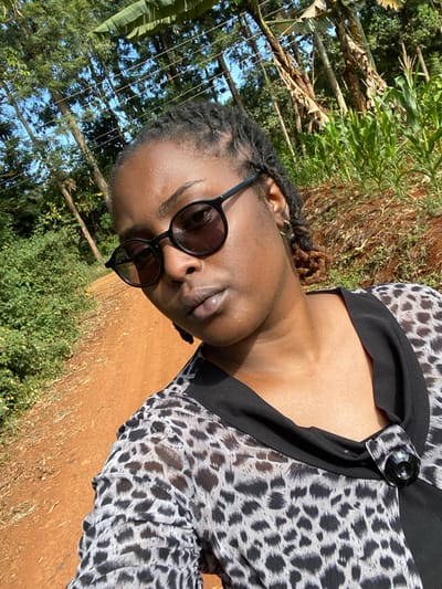A woman with dark skin and dreadlocks wears round, black sunglasses. She is dressed in a patterned blouse with a black collar and large button. The background features green trees and vegetation. The scene appears to be outdoors, with possible corn stalks visible in the distance. The image is taken at an angle, with the ground appearing reddish-brown. The lighting is bright and sunny.