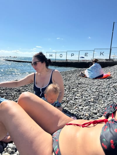 The image depicts a pebble beach in Adlerskiy, Russia, on a sunny day. In the foreground, a person's legs are visible, wearing a bikini with a cherry pattern. To the left, a woman in a dark blue swimsuit and sunglasses is interacting with a baby. The baby is wearing a patterned outfit. In the background, a person wearing a hat and a light blue wrap is sitting on an orange mat, looking at their phone. There is a fence with large letters spelling out "M O P E K" in the background, possibly indicating a location name. The sea is visible with gentle waves and sunlight reflecting on the water. Additional beach umbrellas and a pier can be seen in the distance. The beach is covered with grey and dark pebbles.