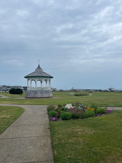 This is a vertically oriented, eye-level shot of a coastal town scene in Oak Bluffs, USA. The image is dominated by a cloudy, overcast sky, suggesting a temperate, possibly humid day. In the foreground, a paved walkway curves through a well-maintained grassy area. To the left of the walkway, a decorative gazebo with a distinctive conical roof and white railings stands prominently. The gazebo appears to be made of wood, with a gray, shingled roof. Beyond the gazebo, the landscape slopes down towards a line of houses, with a distant view of the ocean and a few boats.
The right side of the image features a vibrant flower bed brimming with a variety of colorful blooms, including purple, pink, yellow, and blue flowers, interspersed with green foliage. Further along the walkway, a line of parked vehicles, including cars and buses, is visible, suggesting a public area or parking lot. The overall impression is of a serene, well-kept public space, likely a park or promenade, popular for leisurely strolls and enjoying the coastal scenery. There is no visible text in the image. The scene evokes a sense of calm and natural beauty, typical of a seaside town.