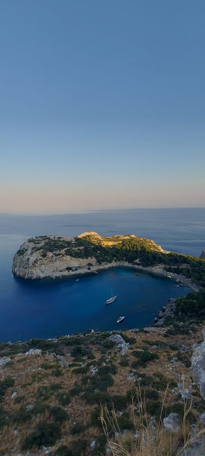 The image captures an elevated view of a picturesque bay in Faliraki, Greece. The bay itself is a deep, calm body of vibrant blue water, enclosed by a rugged, rocky island covered in lush green vegetation. Sunlight illuminates parts of the island, casting shadows and highlighting its textured surface.
Several boats are visible in the water. A prominent sailboat with a white hull and dark mast is anchored in the center of the bay. A smaller, modern white boat is also present. In the distance, the vast expanse of the Mediterranean Sea stretches towards a clear, pale blue sky that transitions to a softer hue near the horizon, suggesting either early morning or late afternoon light.
The foreground of the image shows a rocky, arid hillside dotted with dry grasses and scattered green shrubs, indicating a Mediterranean climate. The overall scene conveys a sense of natural beauty and tranquility, characteristic of the Greek coastline.
