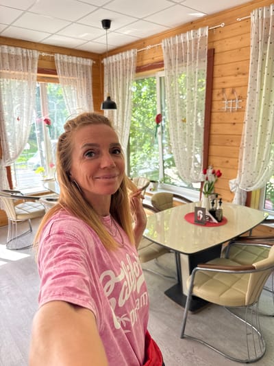 A blonde woman in a pink t-shirt takes a selfie in a cafe. The cafe has wooden walls, white curtains with polka dots, and a tiled ceiling with a black pendant light. There are tables and chairs, with a vase of red tulips on one table. Outside the window, there are trees. The woman is smiling at the camera, and her hair is pulled back. She is wearing a necklace. The overall scene suggests a casual, sunny day in a restaurant or cafe in Serovo, Russia.