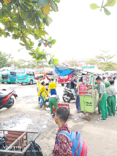 A busy street scene unfolds with children in school uniforms gathered around a green food cart with "TELOR BALADO" written on its side. A man on a motorcycle interacts with the vendor. Several minibuses and motorcycles are parked nearby. A boy with a backpack stands in the foreground, looking towards the crowd. Lush green foliage hangs from above, framing the scene, and waste bins sit on the side. The day appears bright, although the timestamp indicates "14 Jan 2026".
