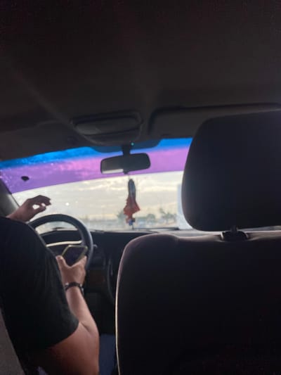 The image depicts the interior of a vehicle. The driver's hands are visible on the steering wheel, with a phone visible in their hand. The windshield shows an exterior scene, including what appears to be a structure, possibly a building. The light through the windshield is refracted, creating a purple and blue effect. A hanging object is suspended from the rearview mirror. The background scene has elements of El Dátil, Venezuela.
